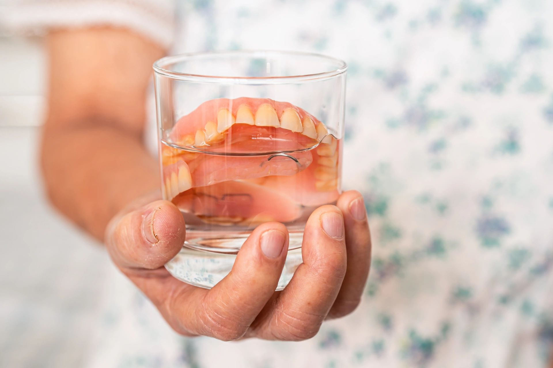 Hand holding a glass with dentures soaking in water, showing the importance of proper denture care and replacement over time.