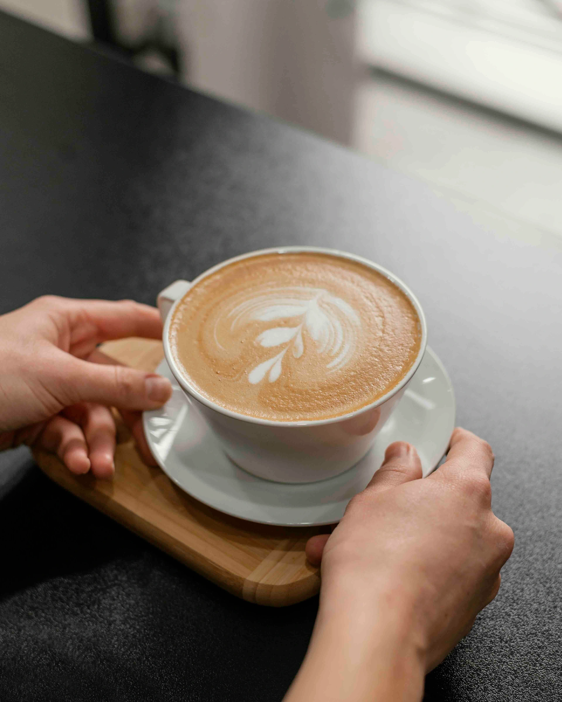 Hands holding a cup of coffee with latte art on a white saucer