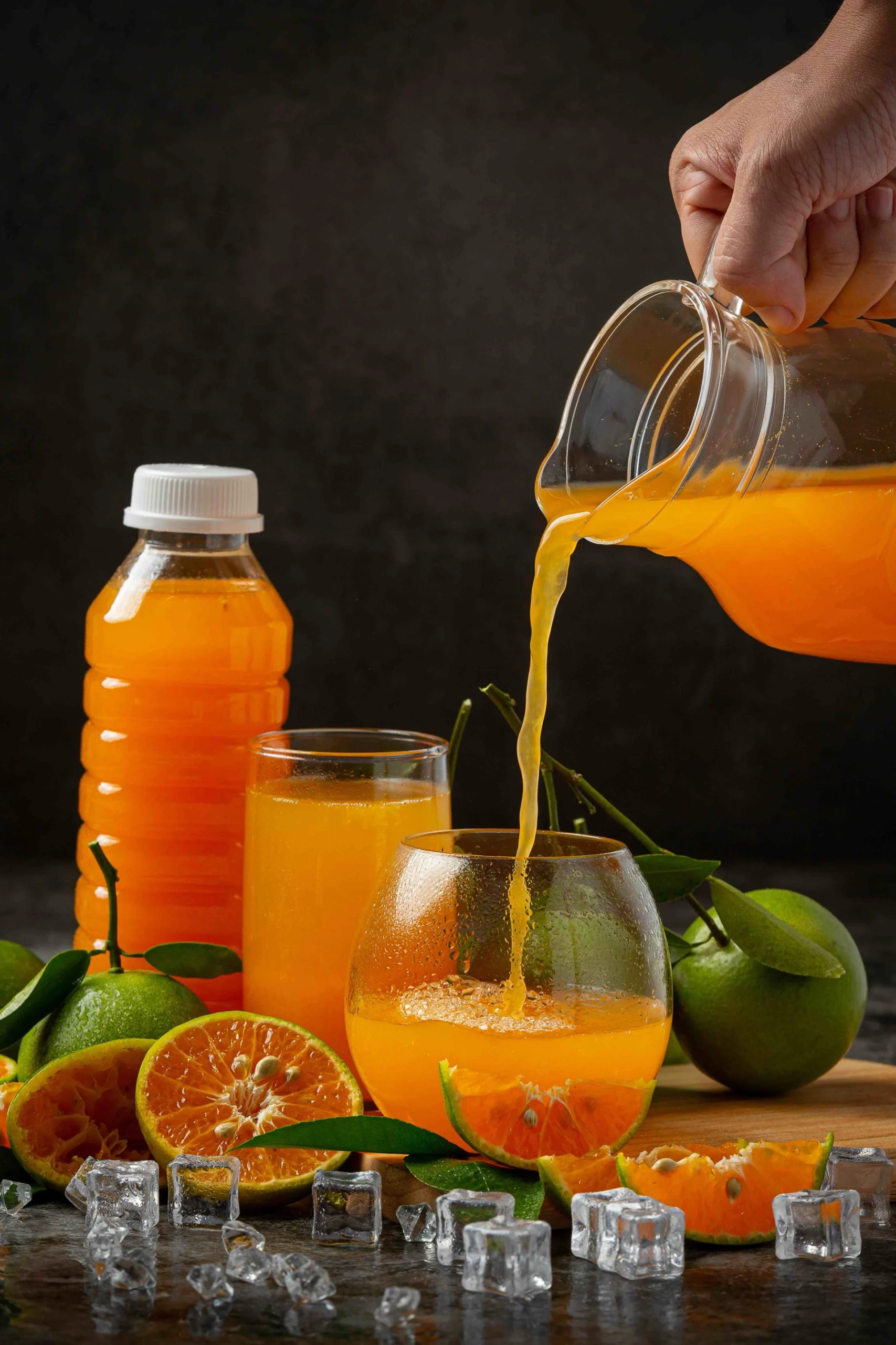 Hand pouring orange juice from a pitcher into a glass with fresh oranges, ice cubes, and bottled juice on a dark background