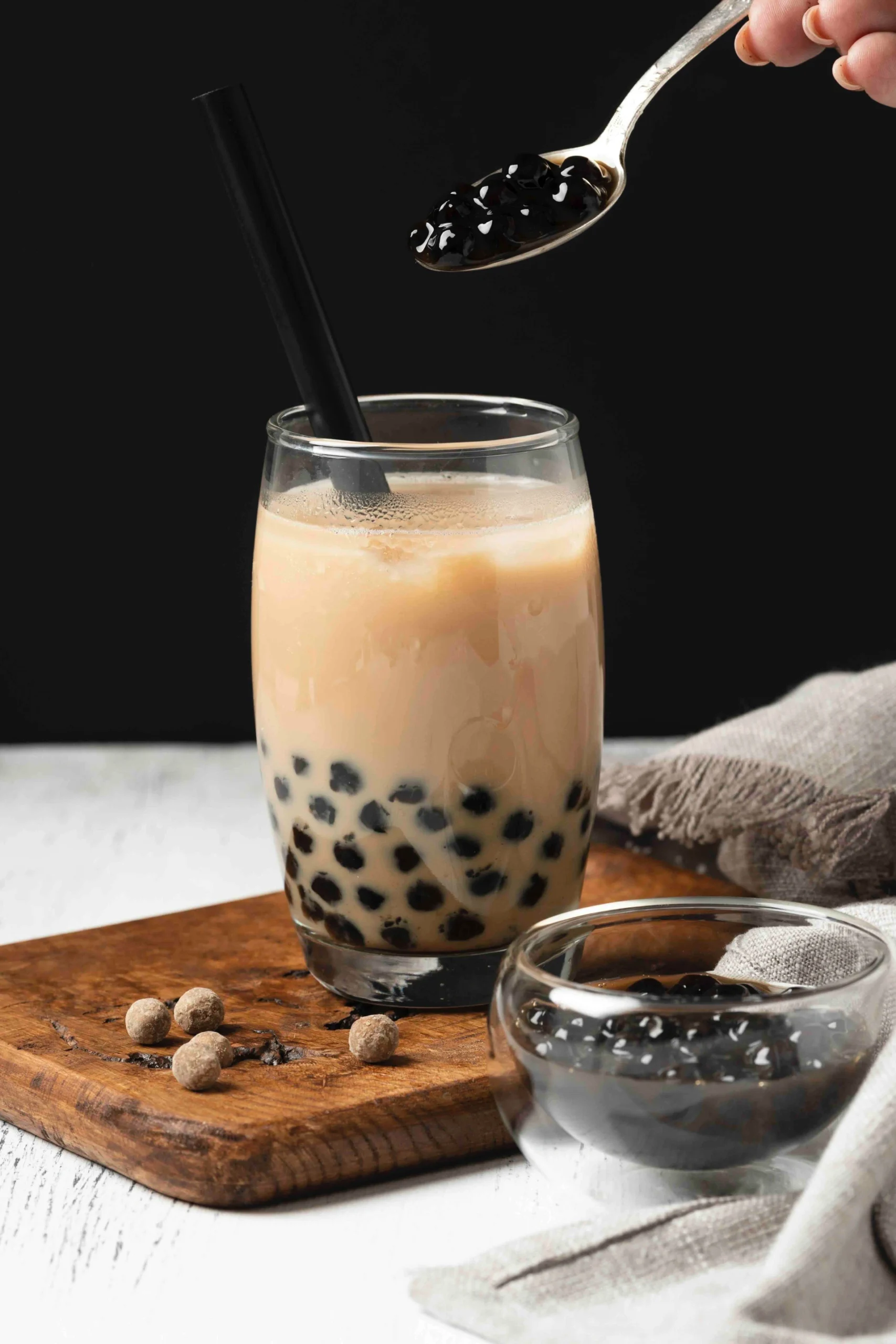 Glass of milk tea with black tapioca pearls and straw on a wooden board, with a spoon adding pearls
