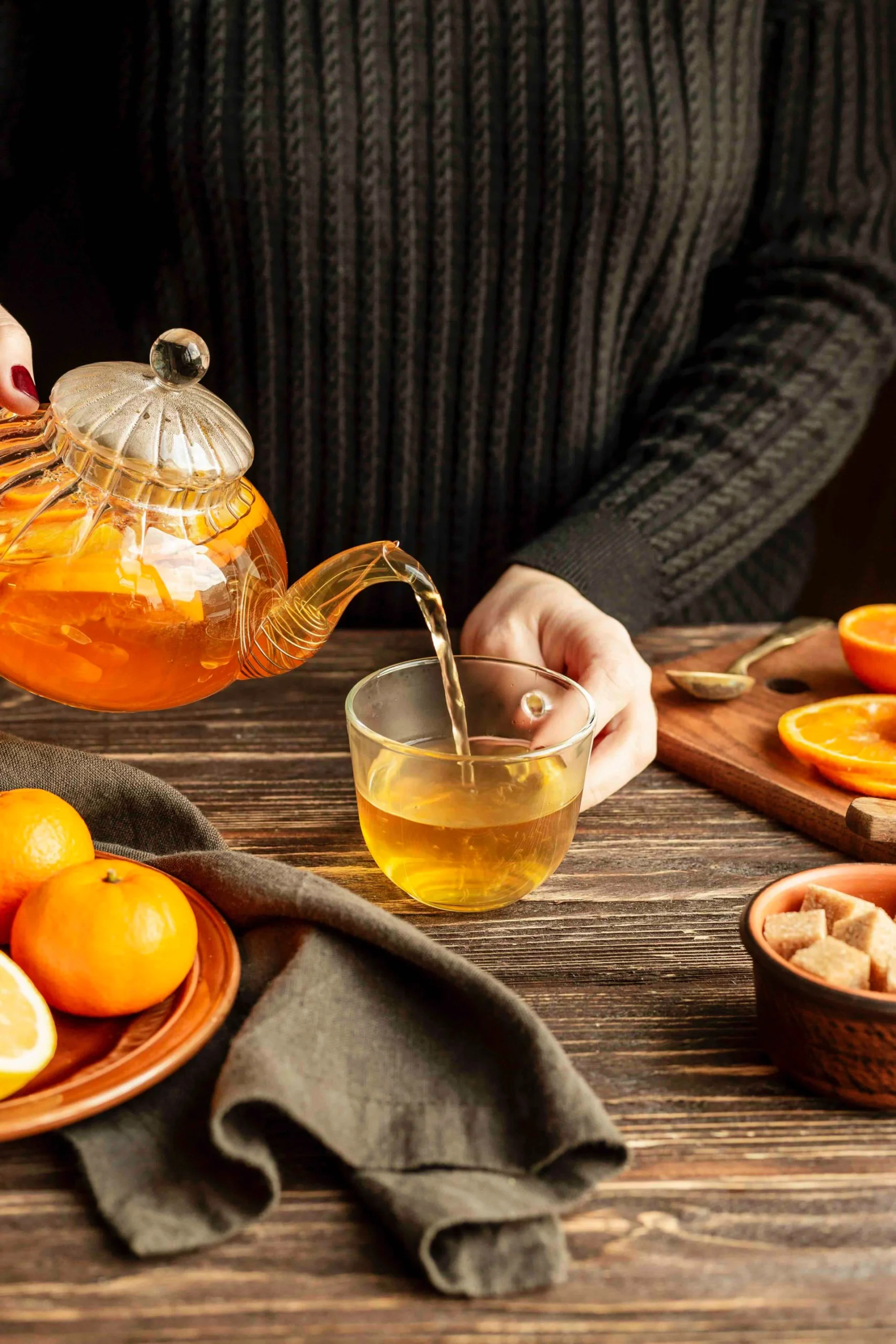 Person pouring freshly brewed tea from a glass teapot into a clear cup beside oranges and sugar cubes
