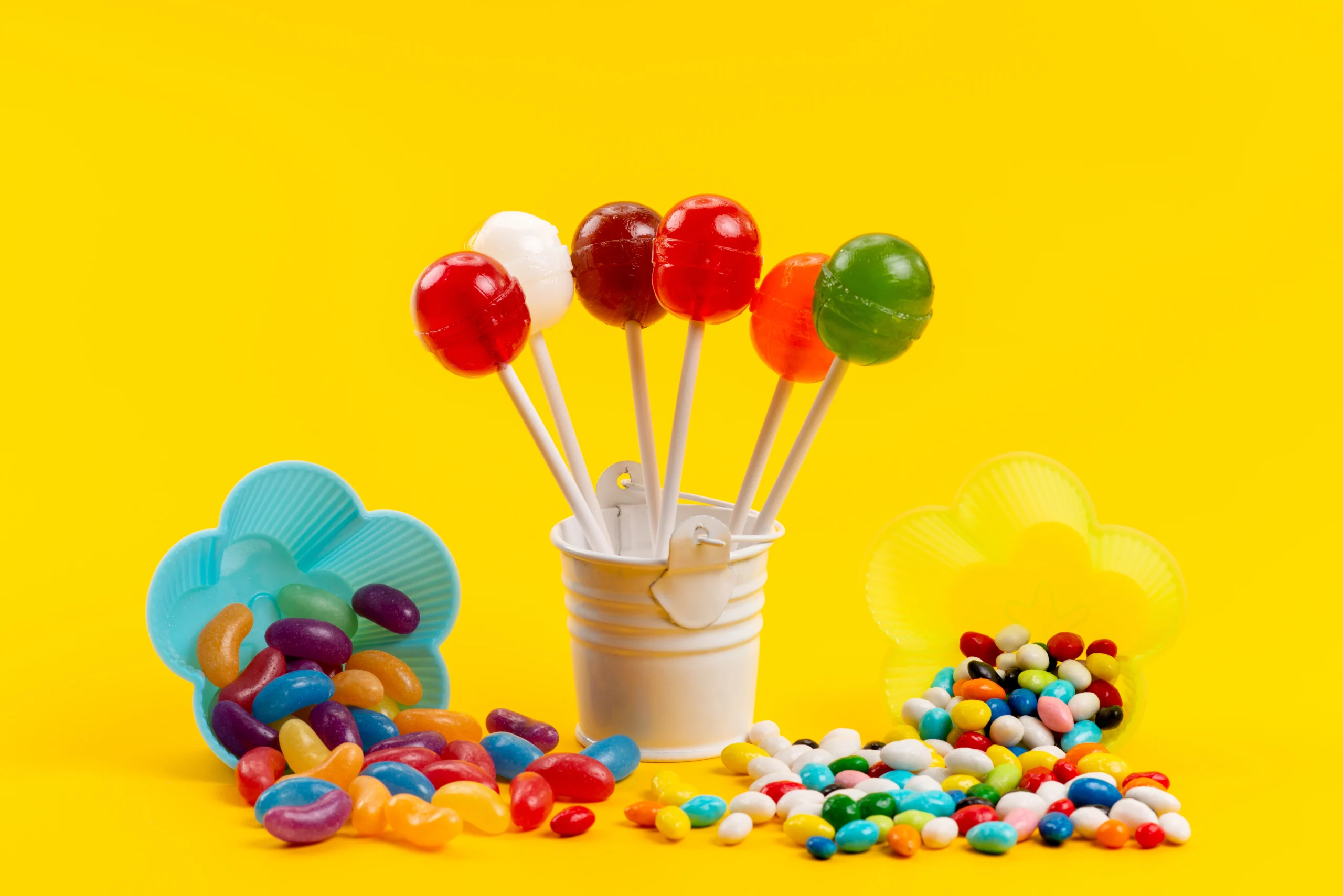 Assorted colorful candies and lollipops on a bright yellow background, representing sugary foods to avoid after teeth whitening.