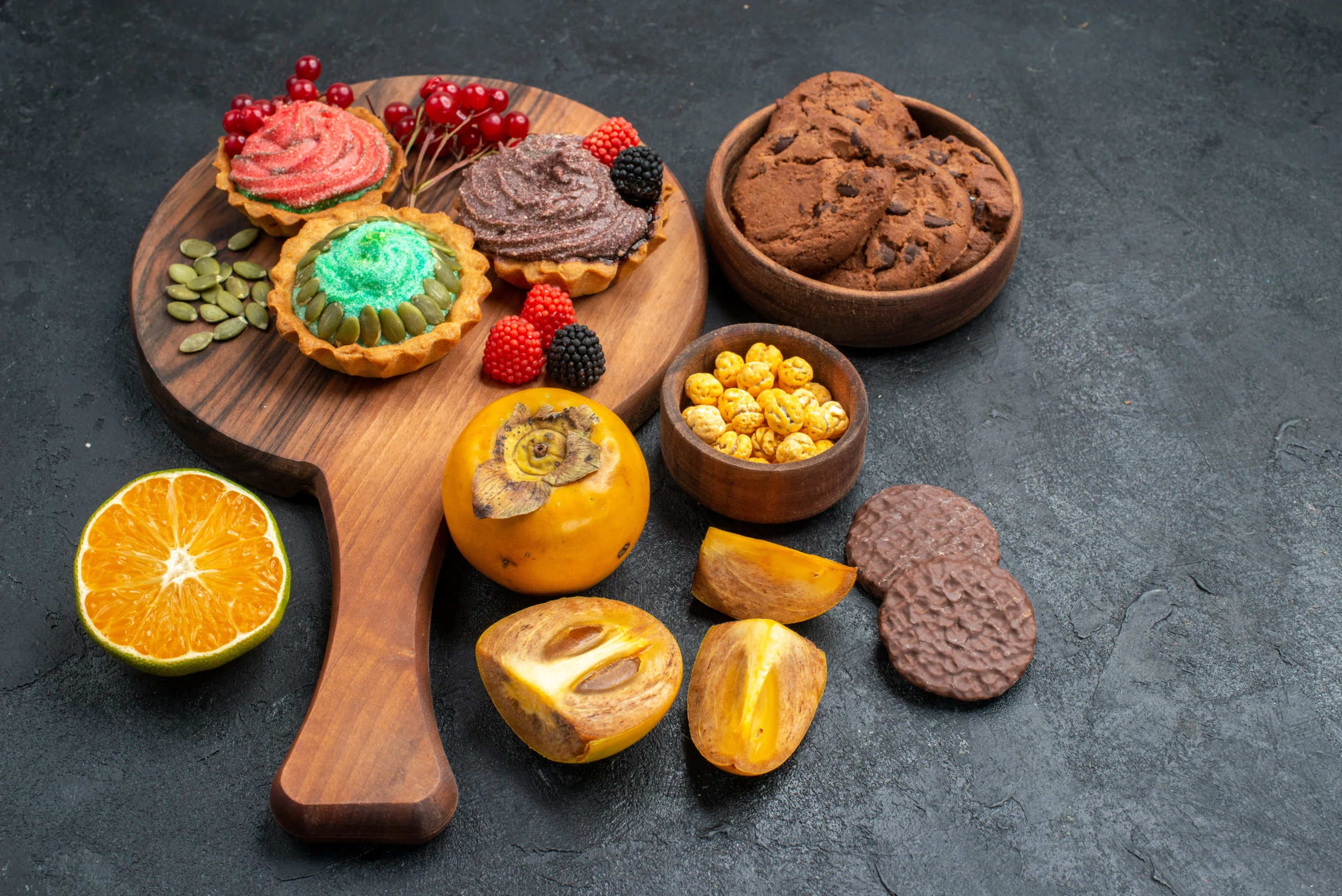 Assorted dark-colored pastries, cookies, and fruits displayed on a wooden board against a dark background.